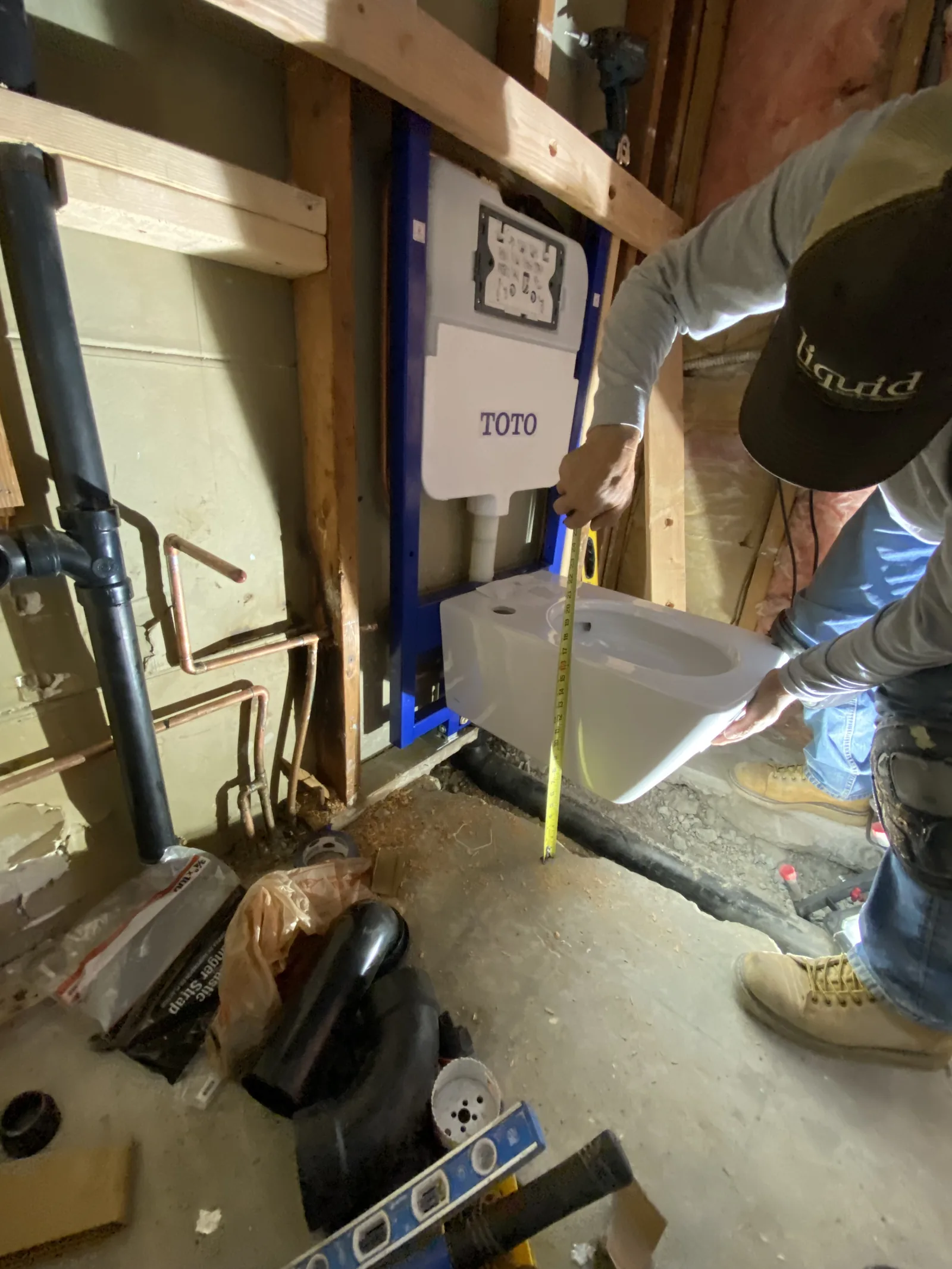 A worker test-fitting a TOTO wall-mounted toilet bowl onto its concealed carrier frame while measuring its height
