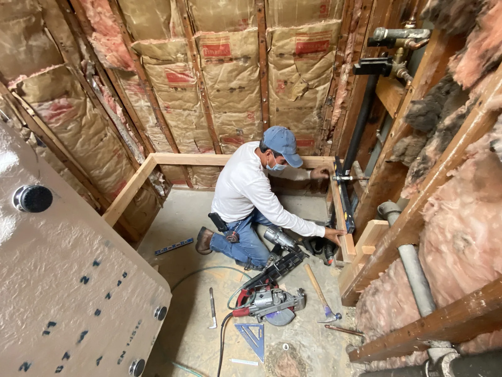 A worker building new stud framing with a nail gun and level inside a gutted bathroom