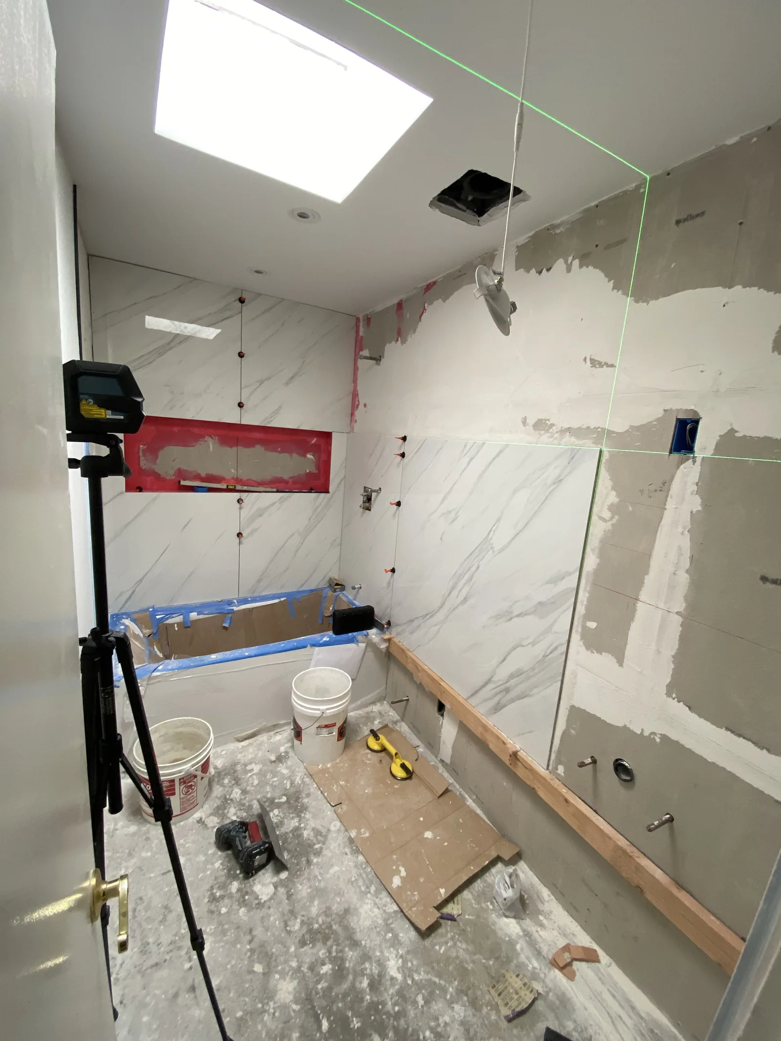 A wide view of a bathroom with tile progressing across two walls, skylight visible above, laser level in use