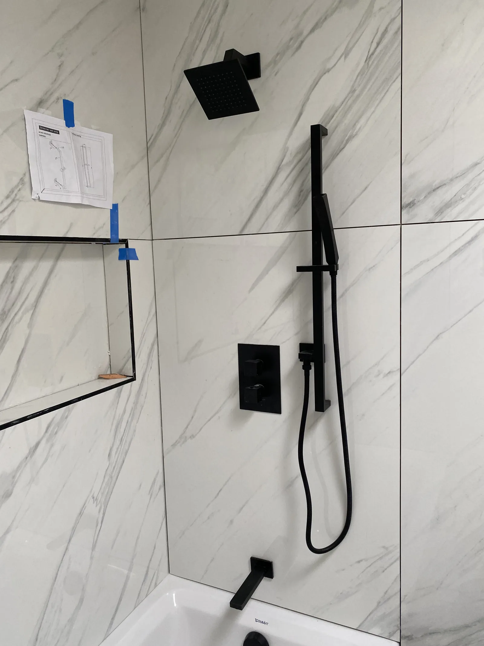 A fully installed matte black shower system with rain head, slide bar, and hand shower, with instructions taped to the wall
