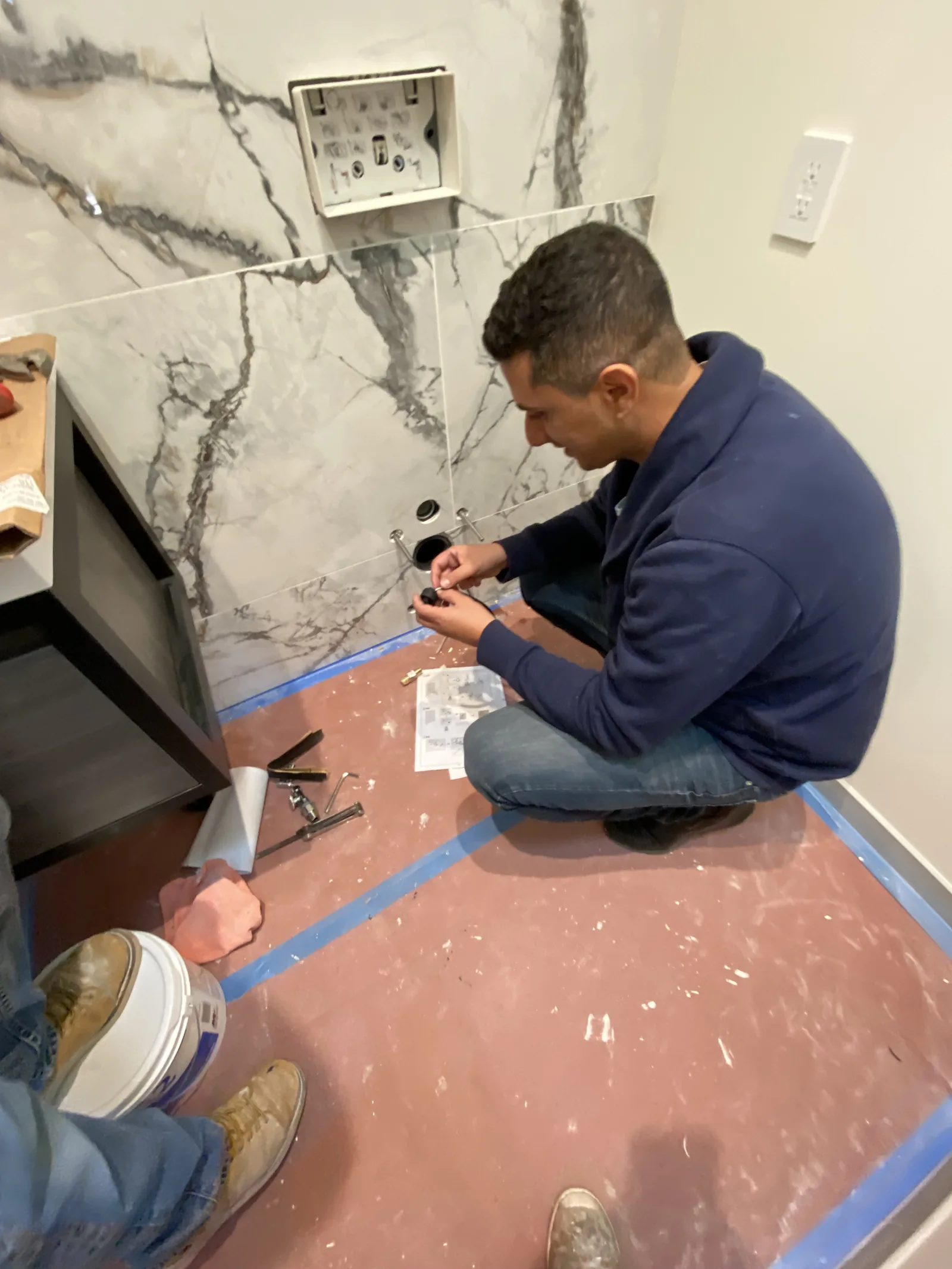 A worker assembling the flush button hardware for a wall-mounted toilet's concealed carrier, crouched on the floor