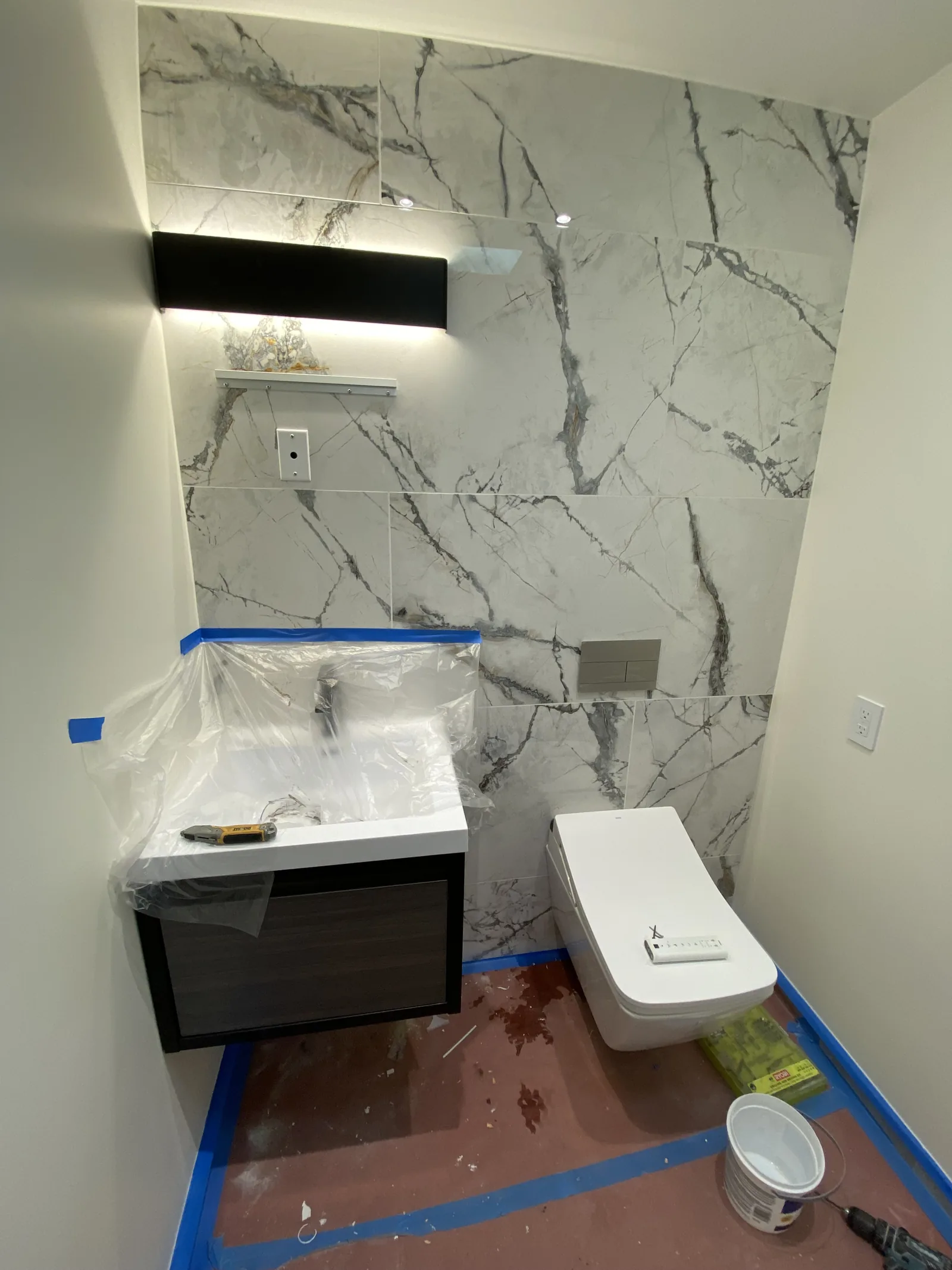 A near-finished powder room with a wrapped vanity, lit mirror, and a toilet with its bidet remote taped to the seat