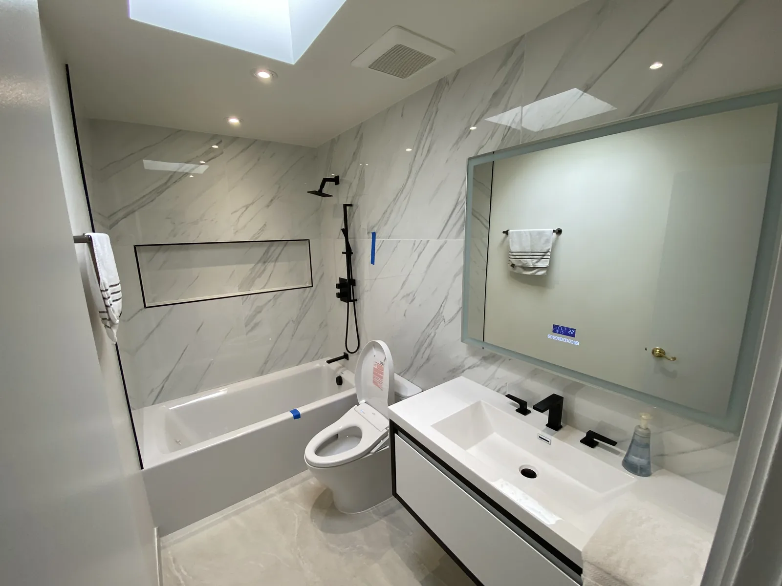 A finished full bathroom with a backlit LED mirror over a floating vanity, black hardware, and marble-look tile