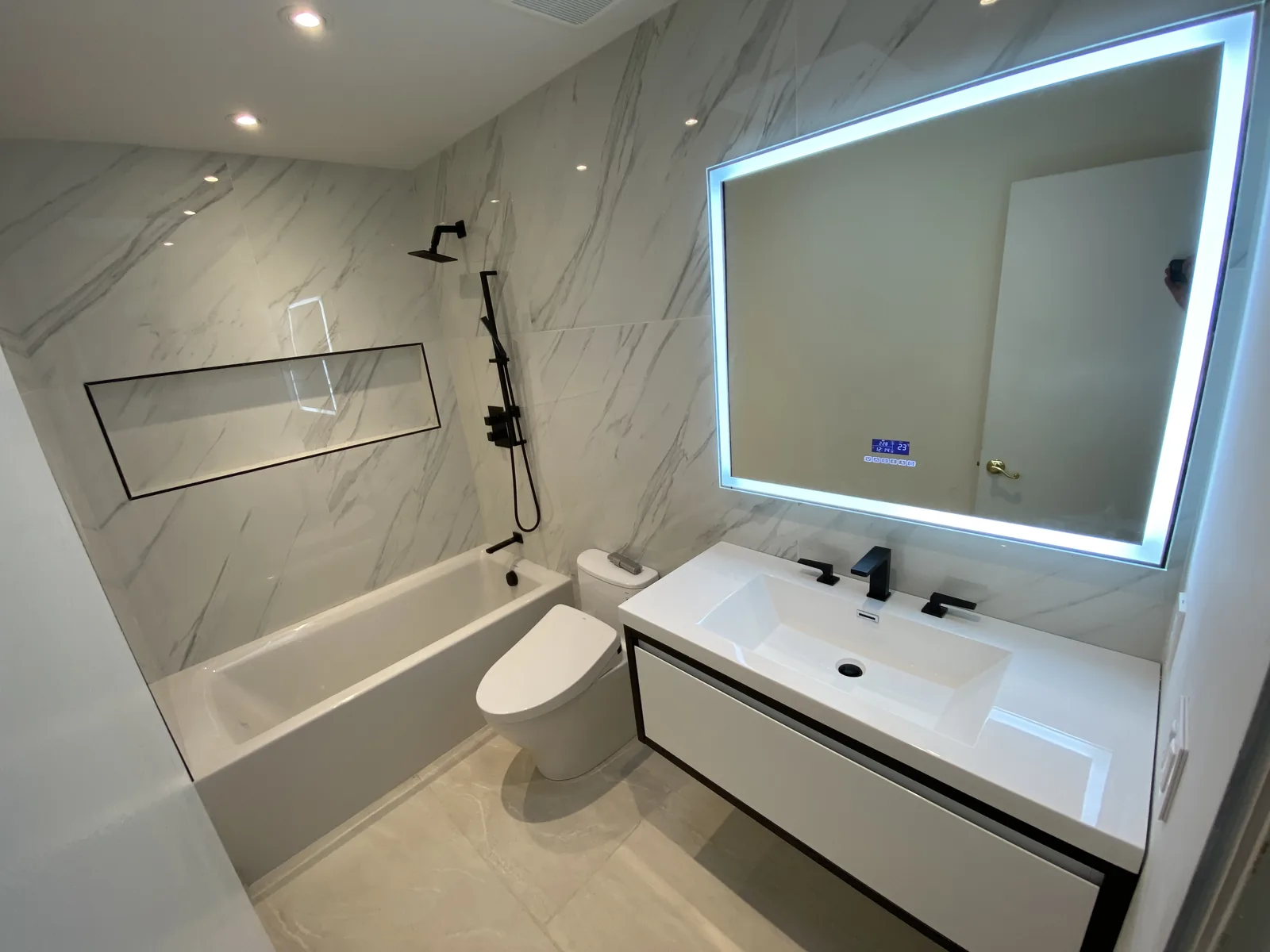 A marble bathroom with a soaking tub, backlit mirror, and black fixtures