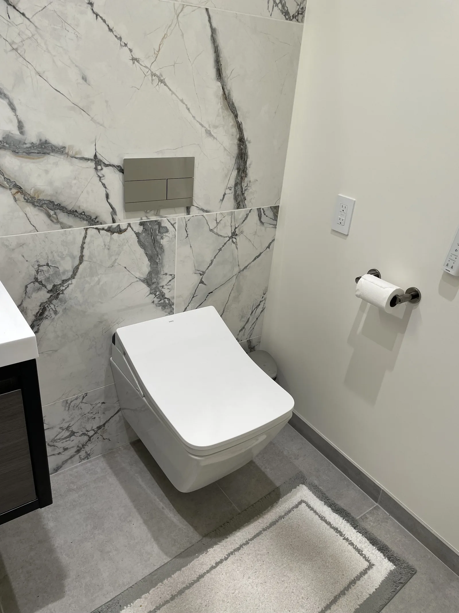 The finished powder room toilet, wall-mounted against a large-format tile wall with a flush plate above it