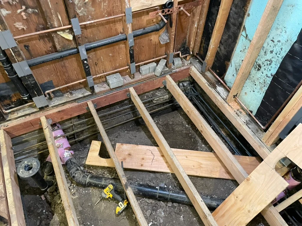 The bathroom floor open with new black drain lines below the joists and rebar along the walls