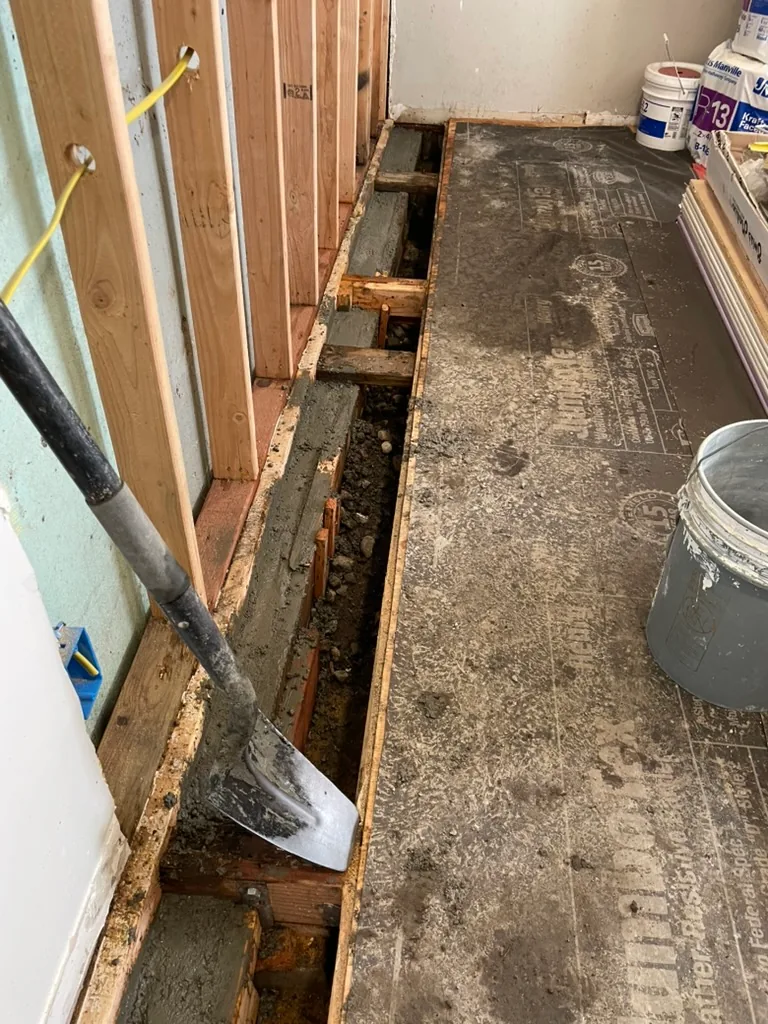 Fresh concrete placed along the base of a wall under the framing, with a shovel
