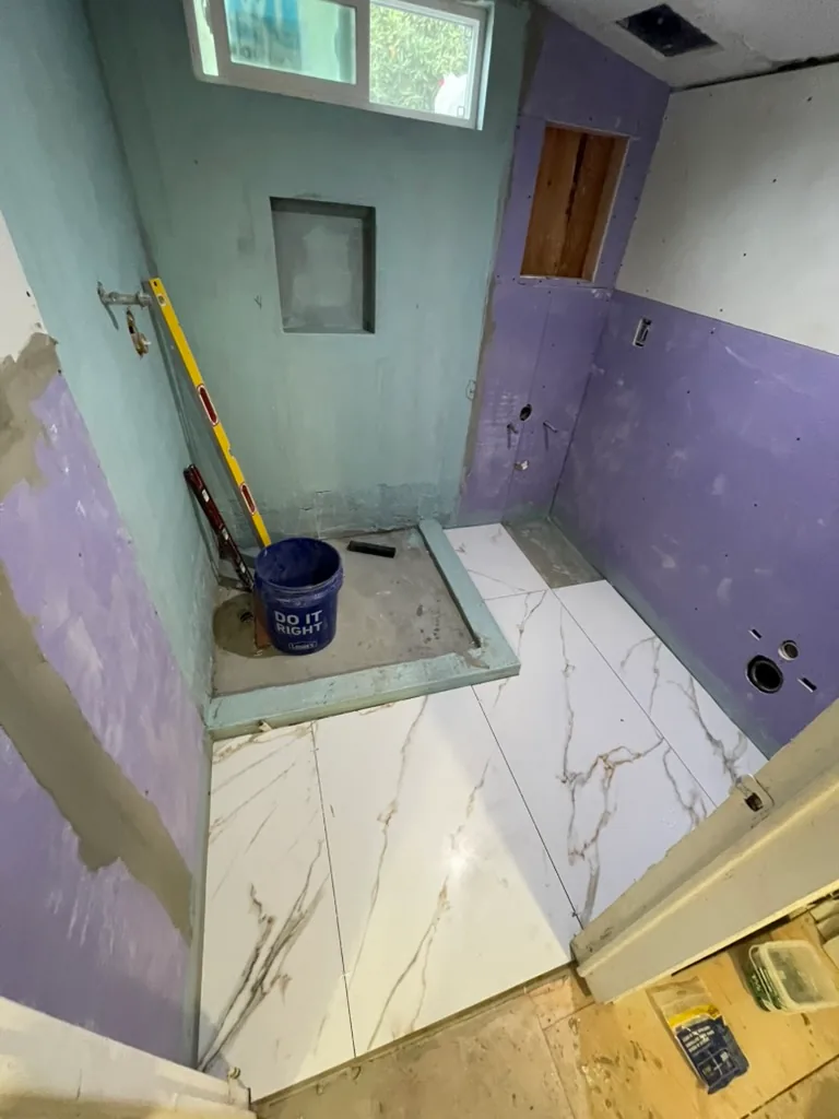 Shower walls coated for waterproofing while large white veined tiles go down on the bathroom floor
