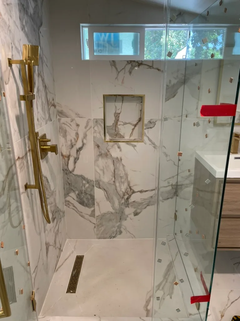 A tiled shower with brass fixtures on a slide bar, a brass-edged niche, a linear drain and frameless glass