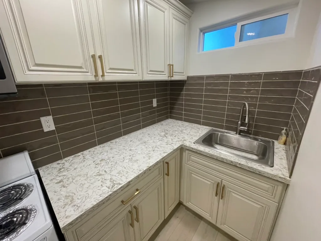 A speckled kitchen counter with a stainless sink, a high-arc faucet, brass pulls and a dark tile backsplash