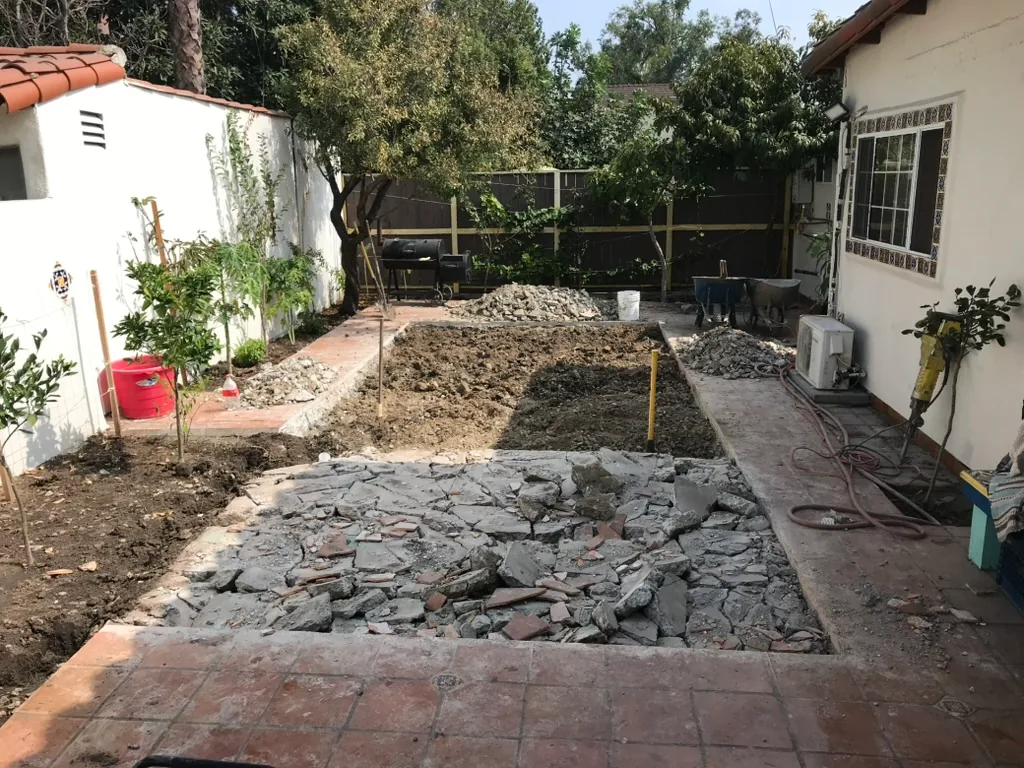 Broken concrete and freshly dug soil in the middle of a terracotta patio, looking toward the back fence