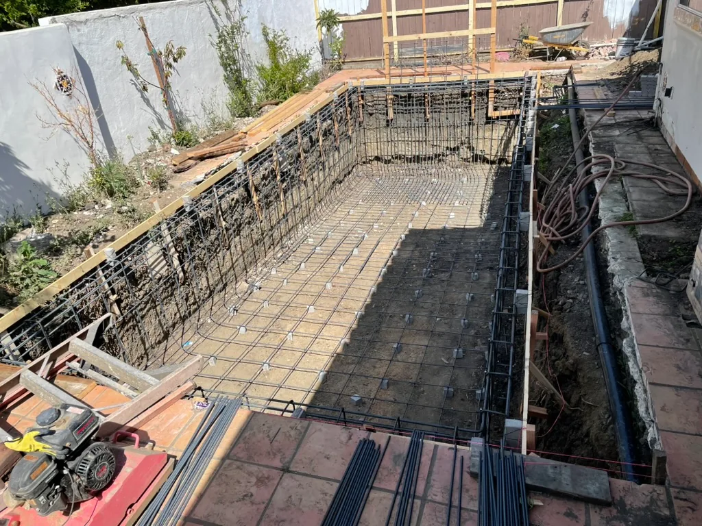 A rebar grid across the pool floor and walls, beside a trench along the house