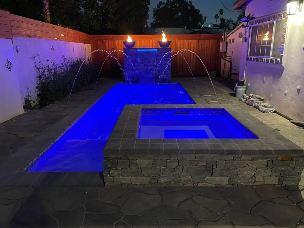 Deck jets arcing into the blue-lit pool at night, with fire bowls lit