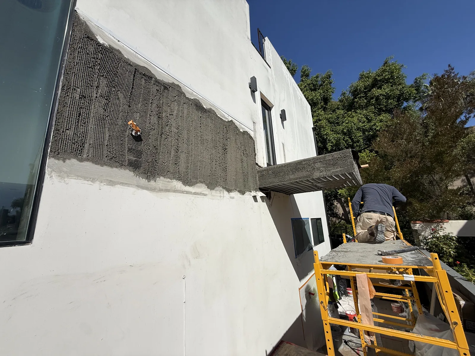 A textured waterproof membrane applied to the exterior wall beneath the balcony, with a worker on scaffold beyond