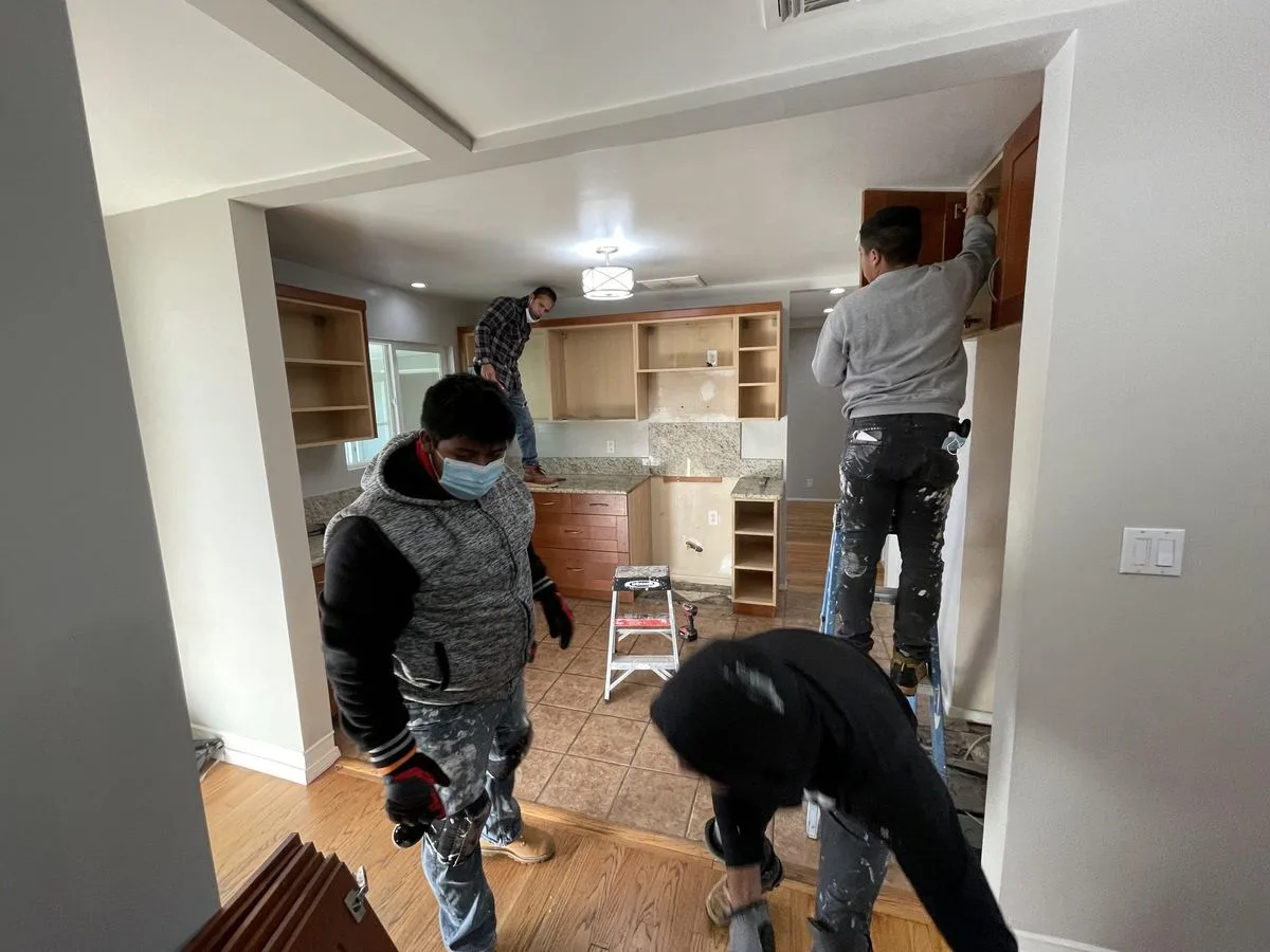 Workers in protective gear in a kitchen with wood cabinetry during renovation