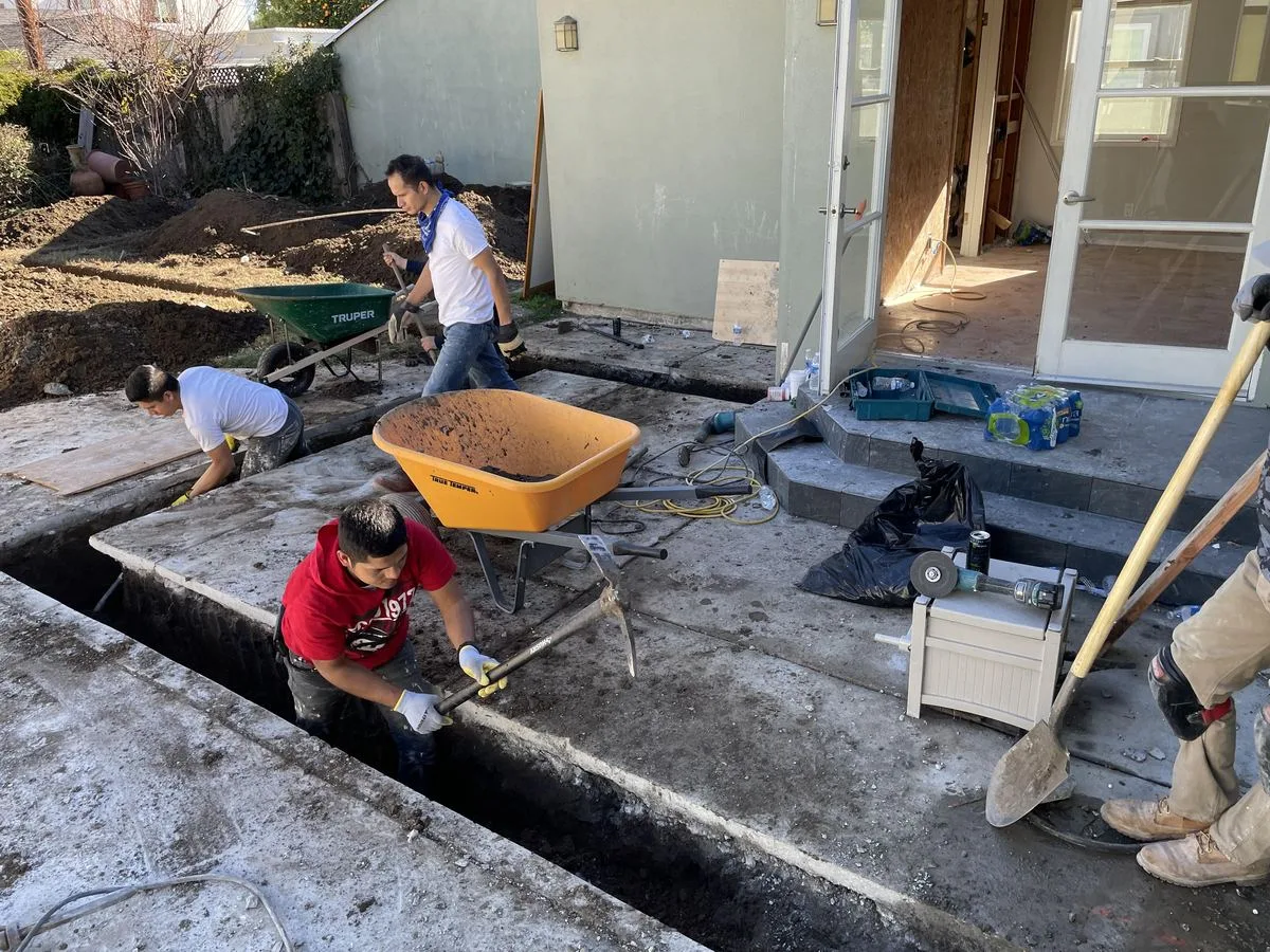 Workers using wheelbarrows to place concrete along a foundation trench in a backyard