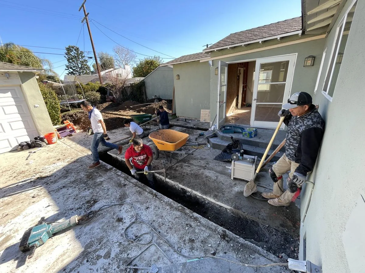 Workers pouring and finishing a concrete foundation slab behind a house, with wheelbarrows and tools