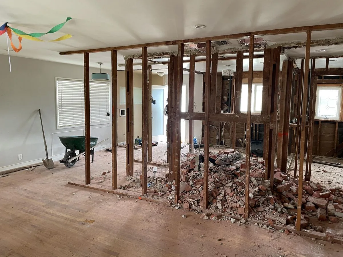 A demolished interior wall down to stud framing, with a wheelbarrow and broom nearby