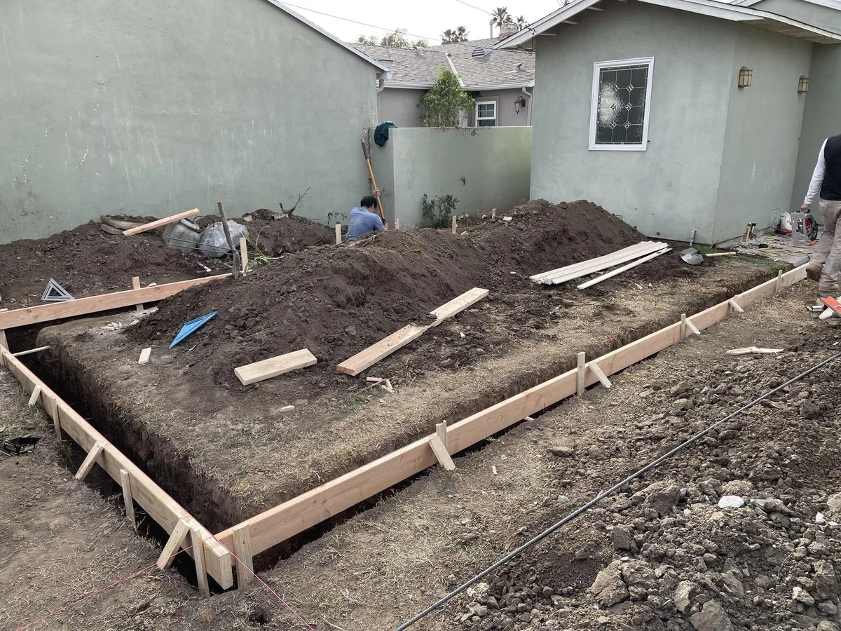 A wood-formed concrete foundation trench with mounded dirt beside it in a backyard