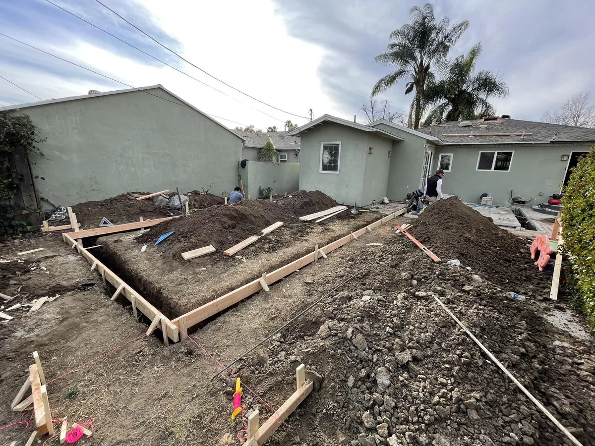 A long wood-formed foundation trench running across a backyard, with dirt piles and a palm tree