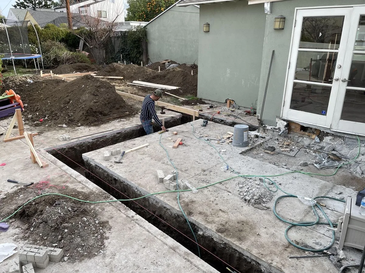 A worker measuring and working at the corner of a foundation trench near a house's back door