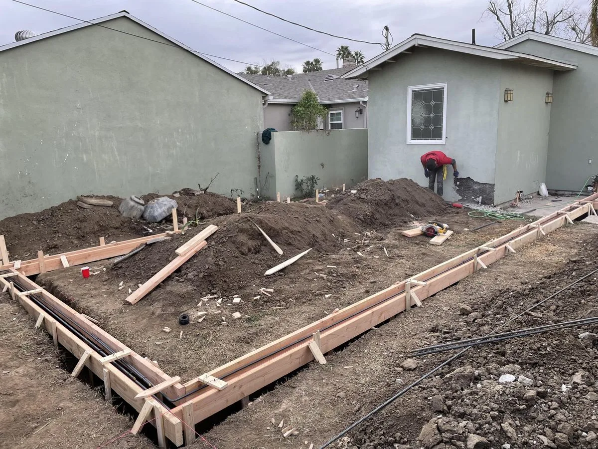 A worker building wood forms for a foundation trench in a backyard, dirt piled nearby
