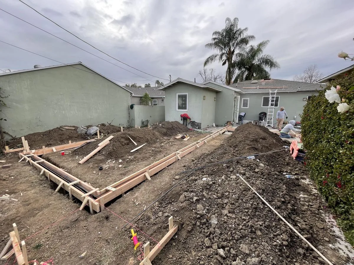 A wood-formed foundation trench under an overcast sky, with a palm tree in the background