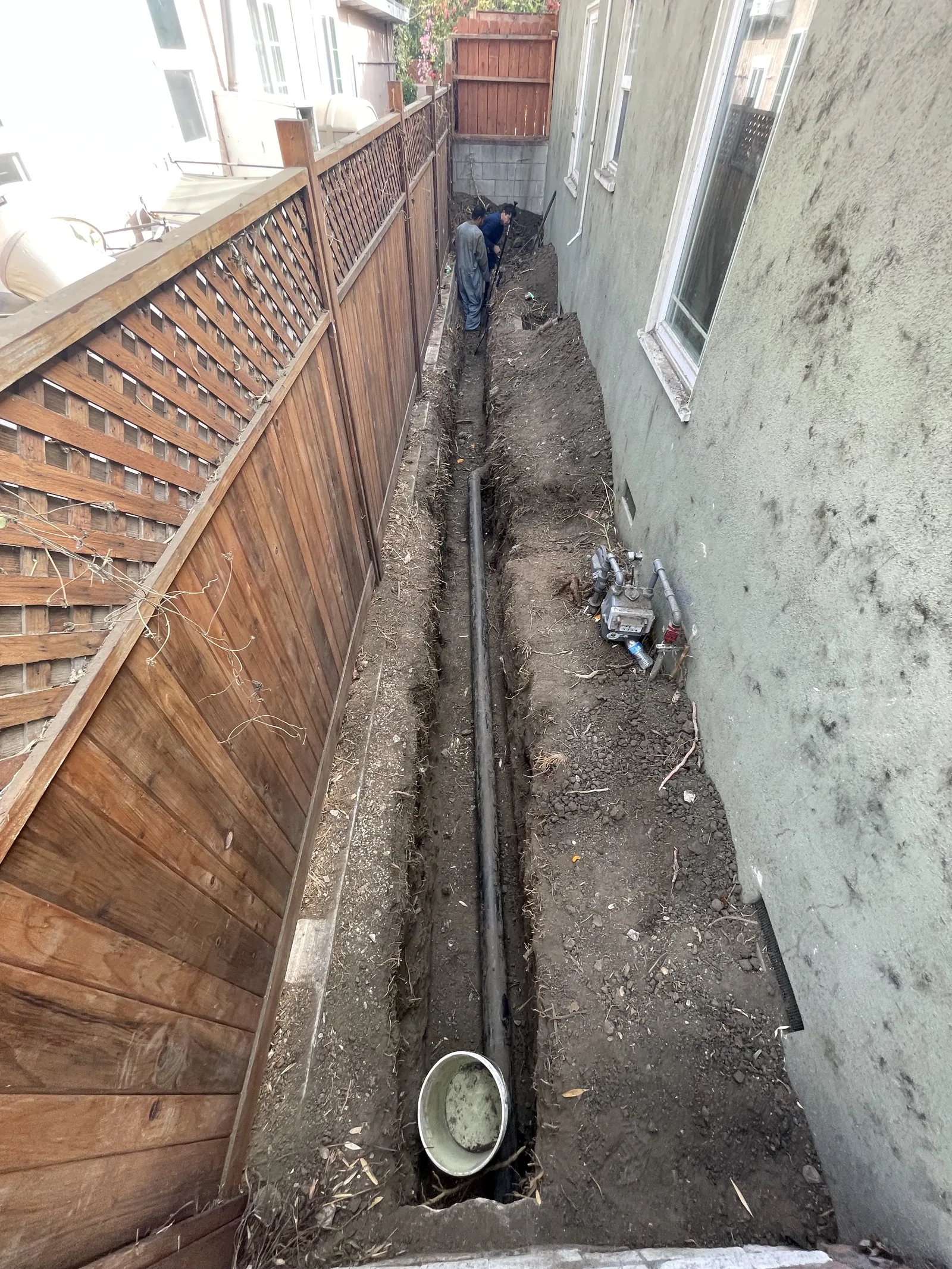 A deep trench running along a fence line, with sewer pipe laid in the bottom