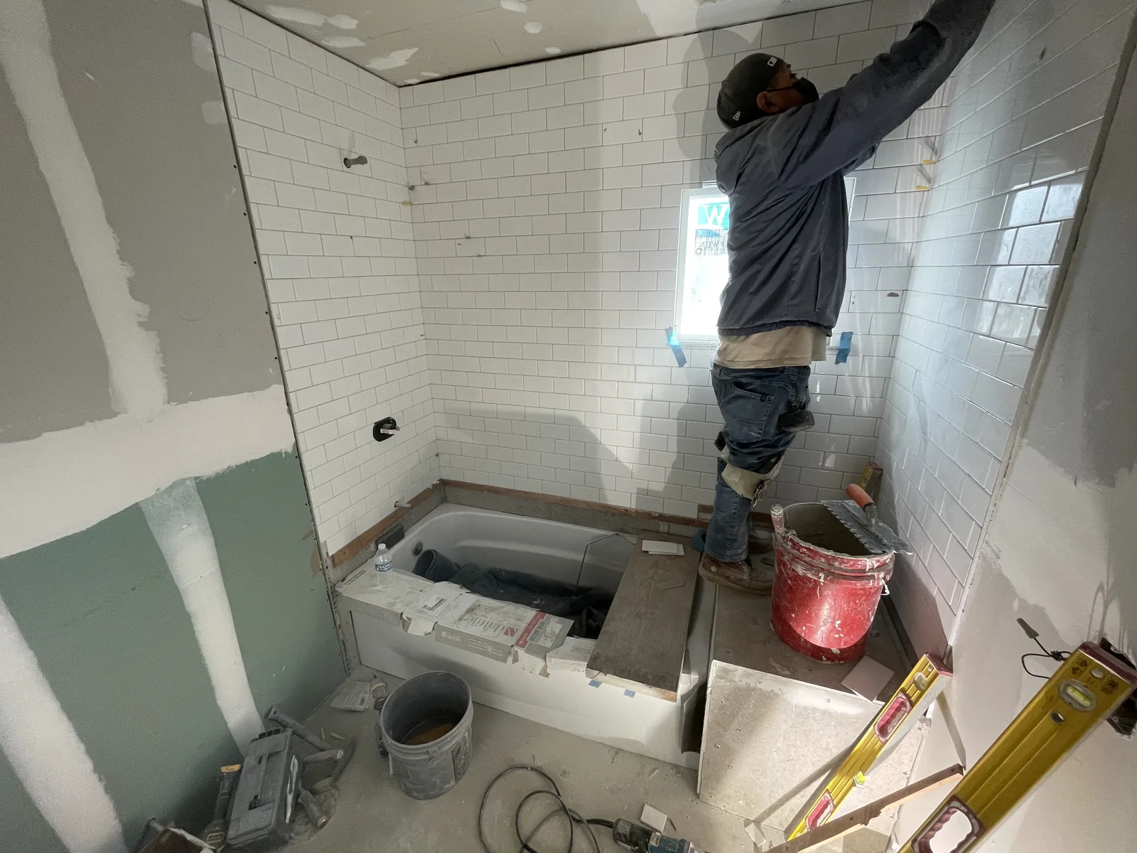 A bathtub covered with protective sheeting, with a worker installing subway tile on the wall above it