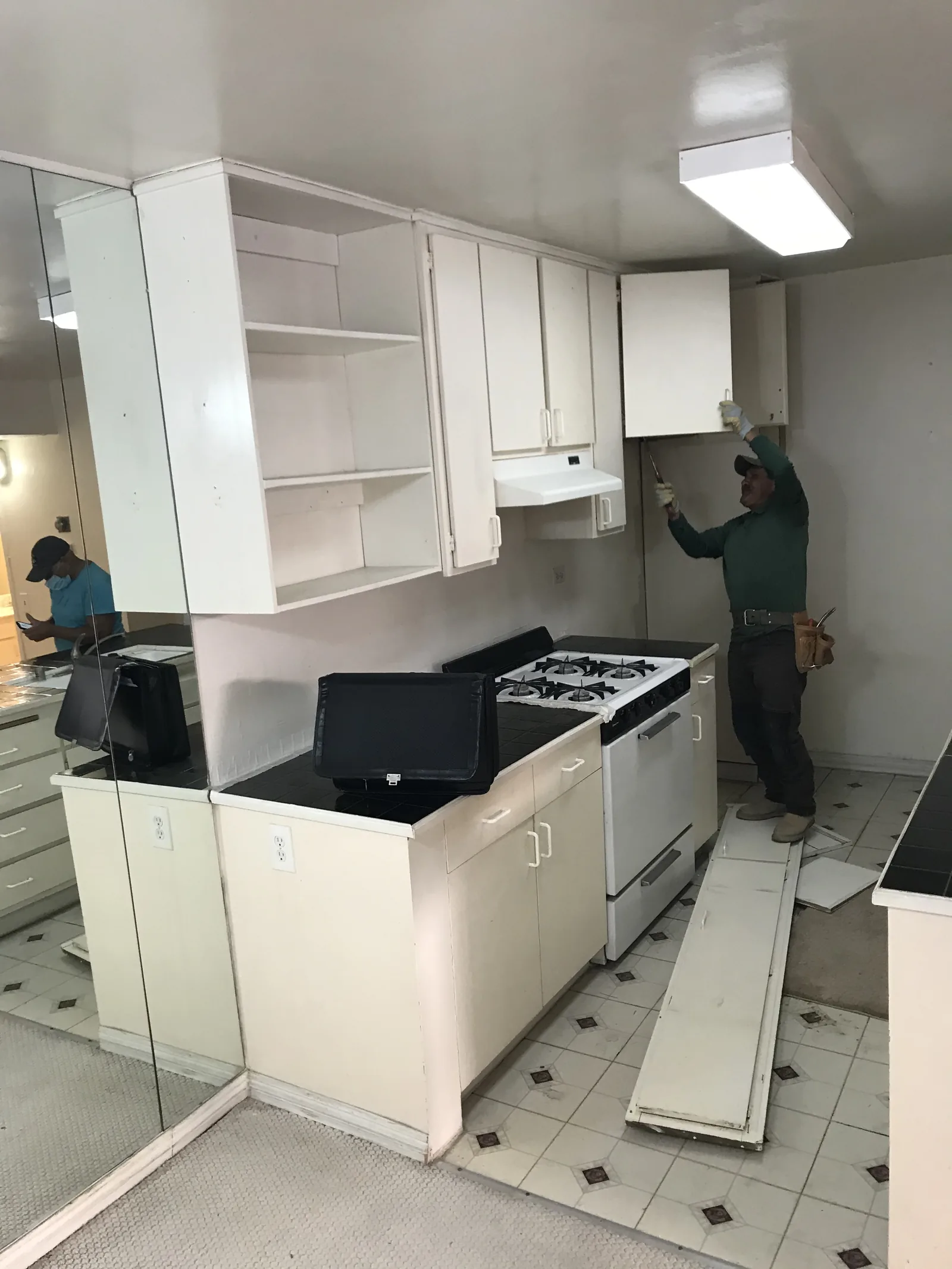 A kitchen mid-demolition with upper cabinets partially removed and a worker installing new upper cabinetry