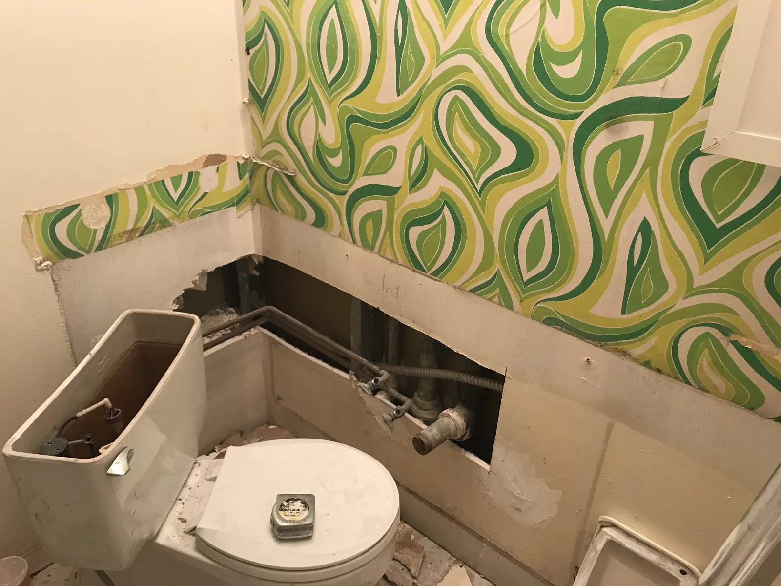 A bathroom mid-demolition with patterned wallpaper partially removed and the toilet disconnected