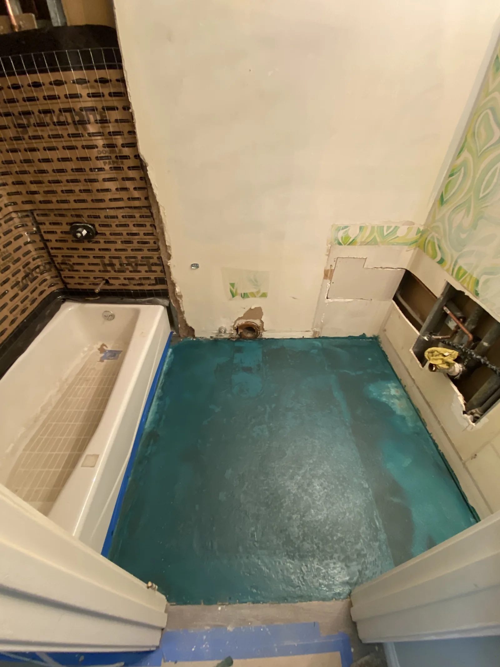 A bathroom floor waterproofed with a teal membrane around the tub, with original wallpaper still on part of the wall